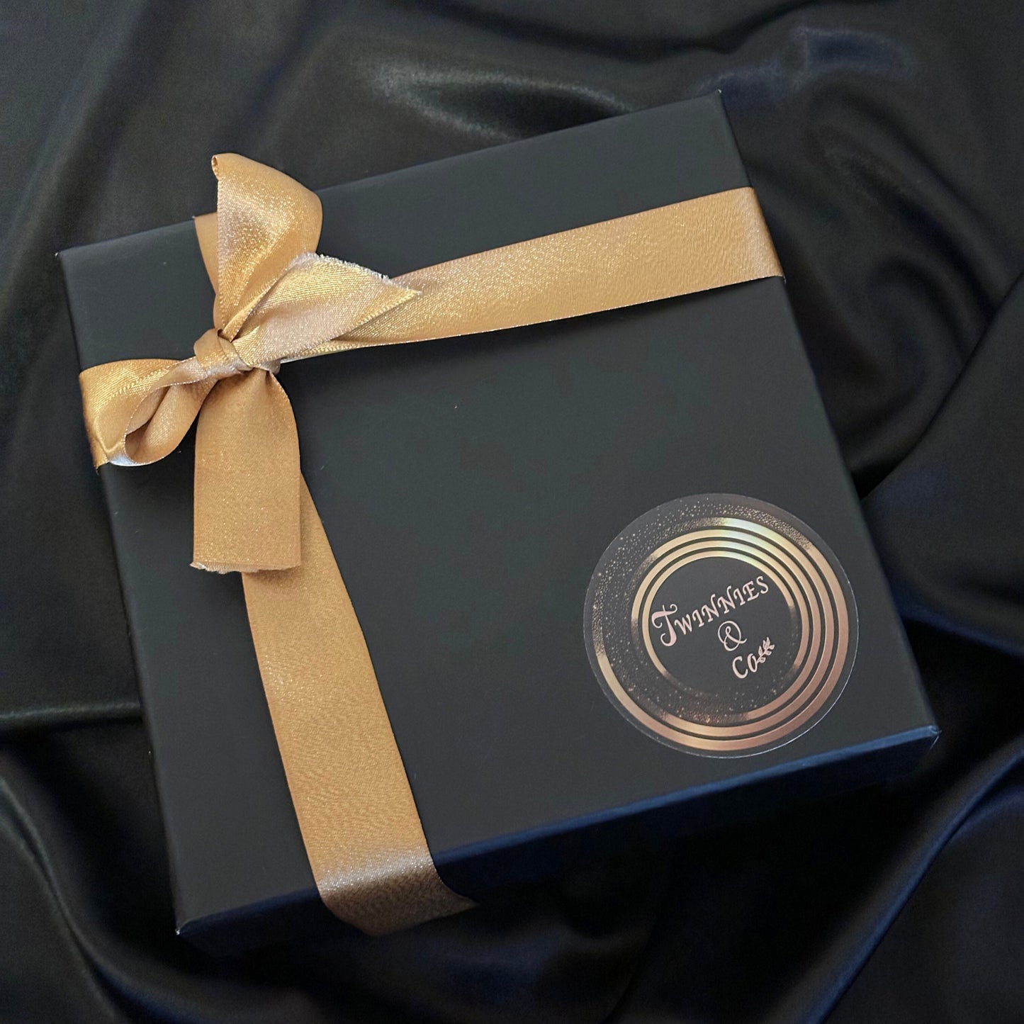 Black gift box with a gold ribbon and decorative label on a dark fabric background