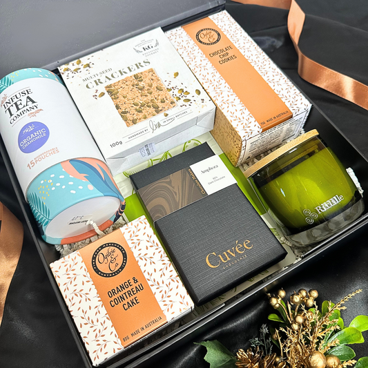 A curated gift box arrangement featuring Cuvée chocolates, an orange & Cointreau cake, crackers, a candle, and a tea , presented in a black gift box with a gold ribbon.