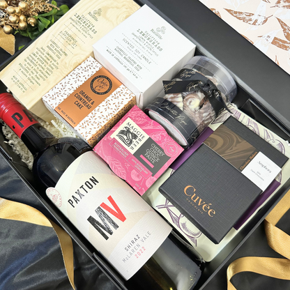 Gourmet food and wine gift hamper featuring premium wine, chocolates, olives, cake, and scented soy candle in elegant packaging.