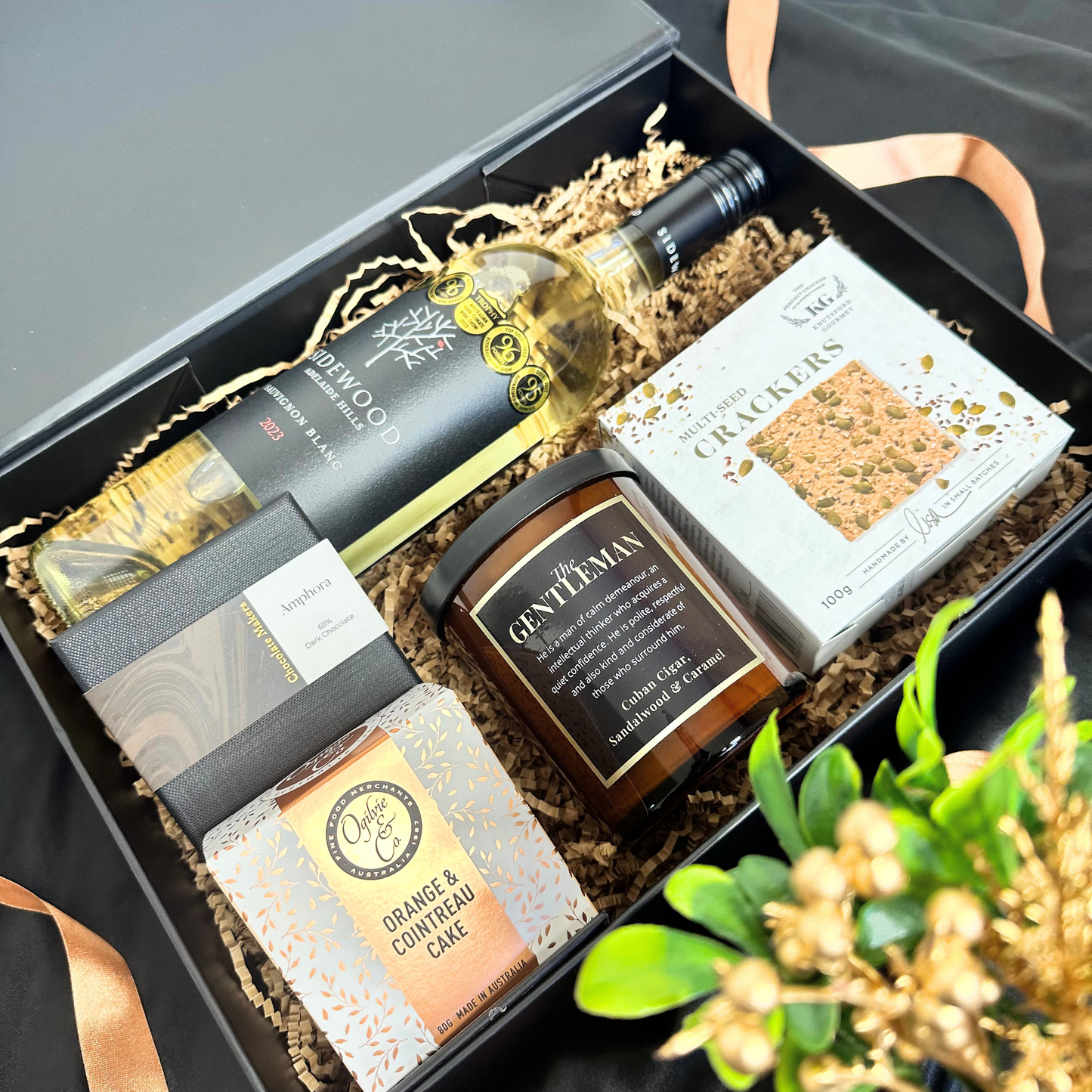 A curated gift box set containing a bottle of Glenwood Shiraz, a candle from Gentleman's, a box of Macadamia Crackers, and an orange and coumphas cake from The Orange Box, arranged in a black gift box with shredded paper filler.
