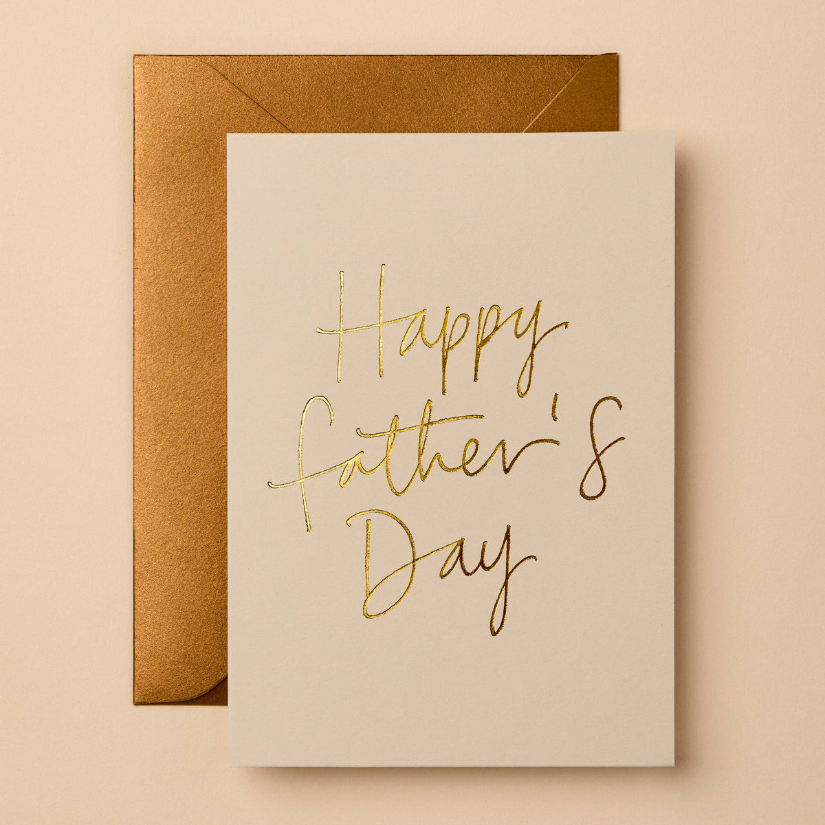 A Father's Day greeting card with elegant gold foil script reading 'Happy Father's Day' on an off-white card, shown with a metallic gold envelope against a warm beige background.